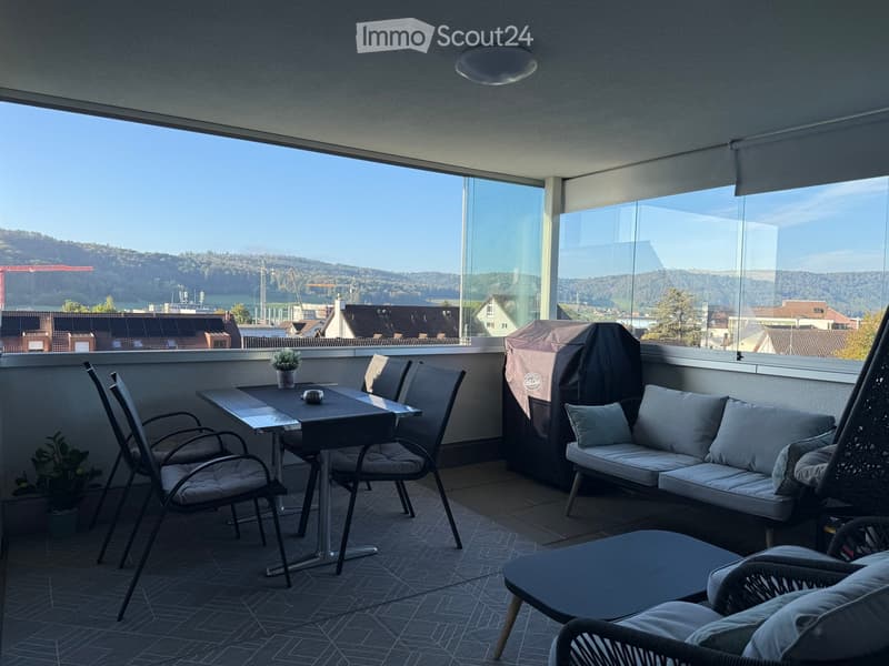 Balcony with large windows offering a scenic view, furnished with a table, chairs, a sofa, and a wicker hanging chair.