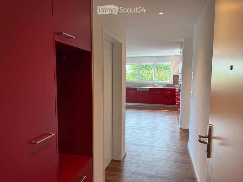 A modern hallway with wooden flooring, red storage cabinetry, and a view into a kitchen area with red cabinets and a window.