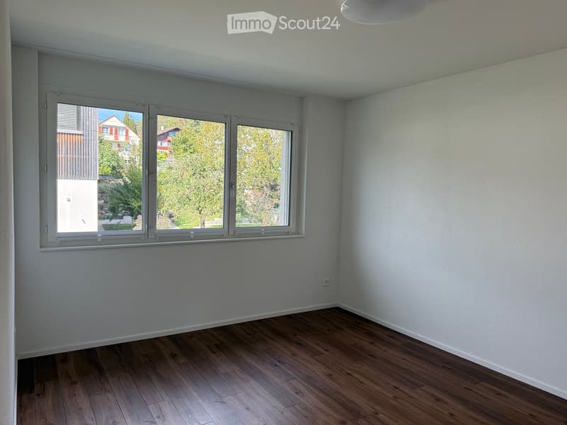 Empty room with light grey walls, wooden flooring, and large windows offering a view of the outside greenery and neighboring houses.
