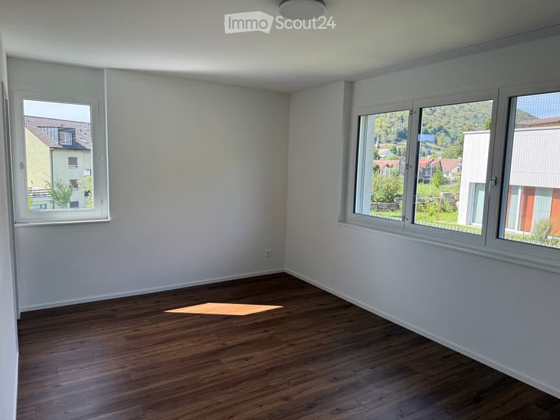 Empty room with wooden flooring, white walls, and large windows offering a view of the surrounding area.