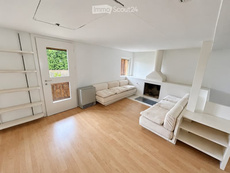 Modern living room with light-colored wooden flooring, white walls, and a fireplace. Features two beige sofas, a white shelf unit, and a small window with a view of greenery.