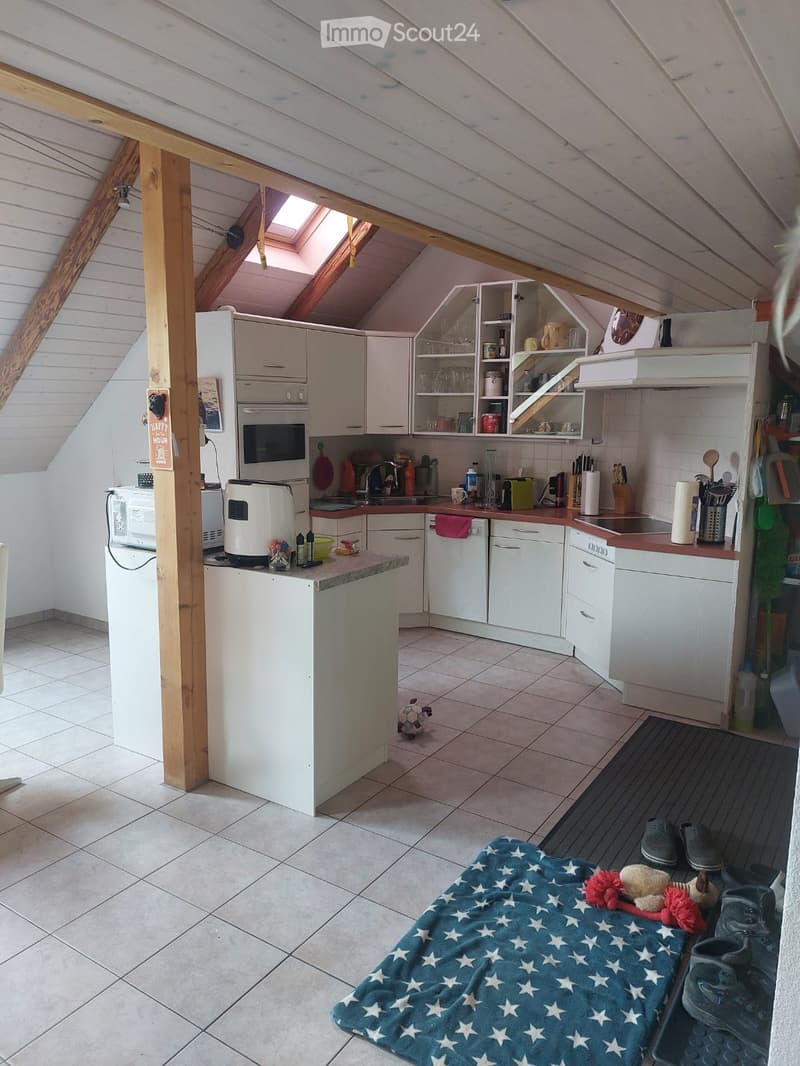 The image shows a cozy attic kitchen with white cabinets, a central island, and various appliances including a refrigerator, oven, microwave, and dishwasher. The space features exposed wooden beams and a skylight, with a small seating area including a blu