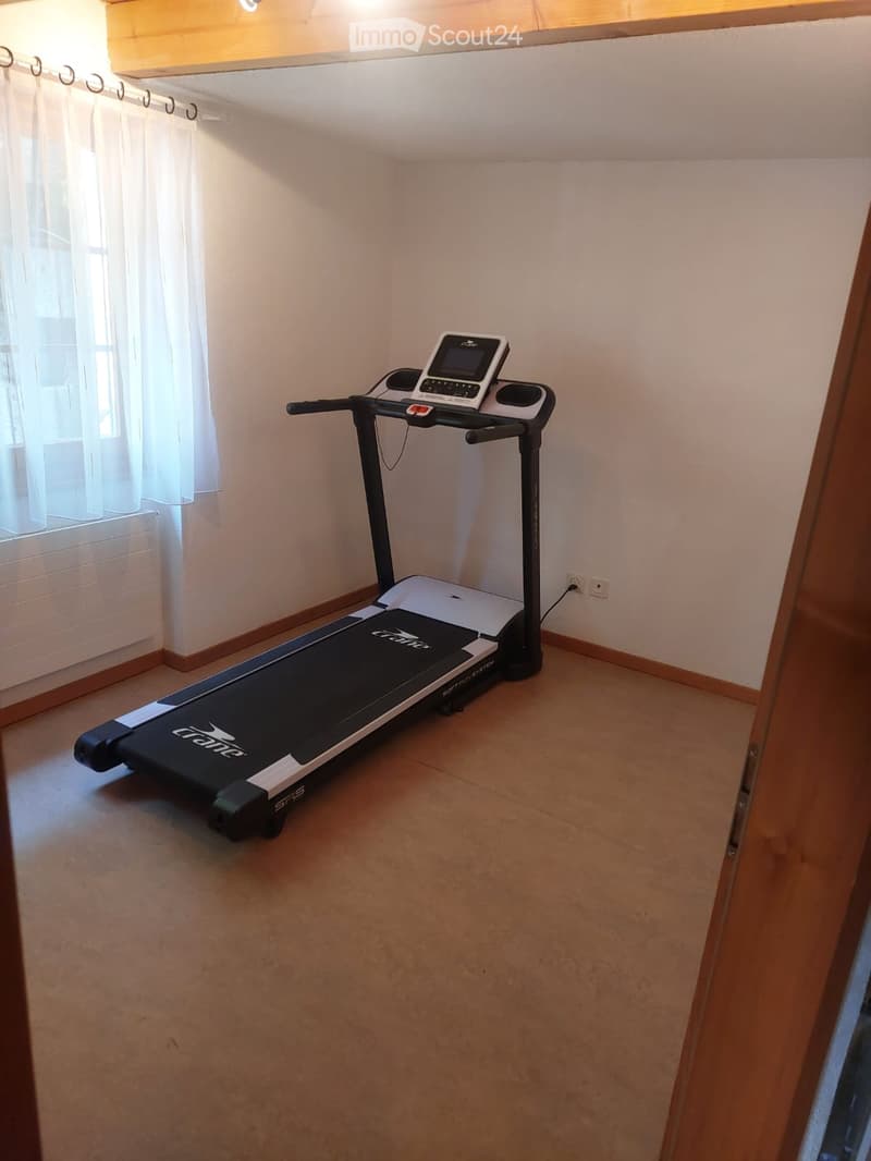 A small, well-lit room with a treadmill positioned near a window with white curtains. The room has light-colored walls and a wooden floor.