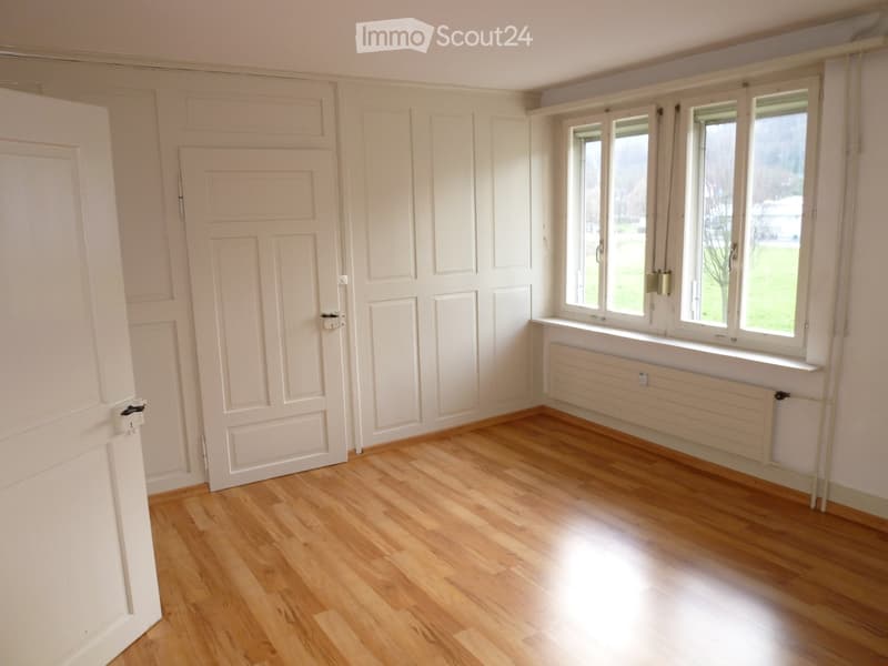 Empty room with wooden flooring, white walls, and multiple windows with radiators underneath. Two white doors are visible on the left side.