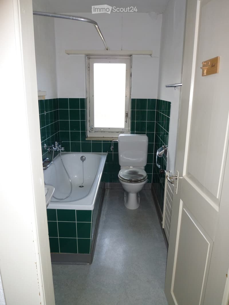 Bathroom with green tiled walls, a bathtub, and a toilet. The room has a window above the bathtub and a door with a towel rack on the side.