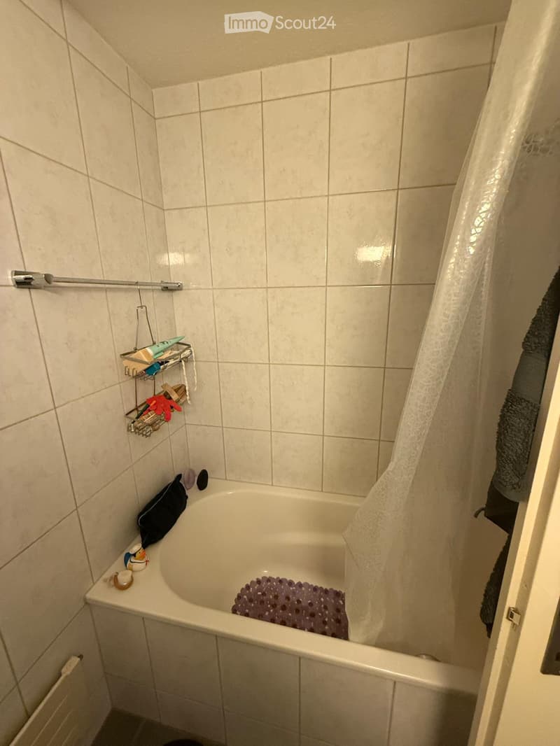 Bathtub with a purple bath mat inside, surrounded by tiled walls and a shower curtain. A towel hangs on the side, and a shelf holds various toiletries.