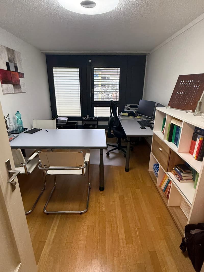 Office space with wooden flooring, two desks, chairs, shelves with books, computer setup, and artwork on the wall.