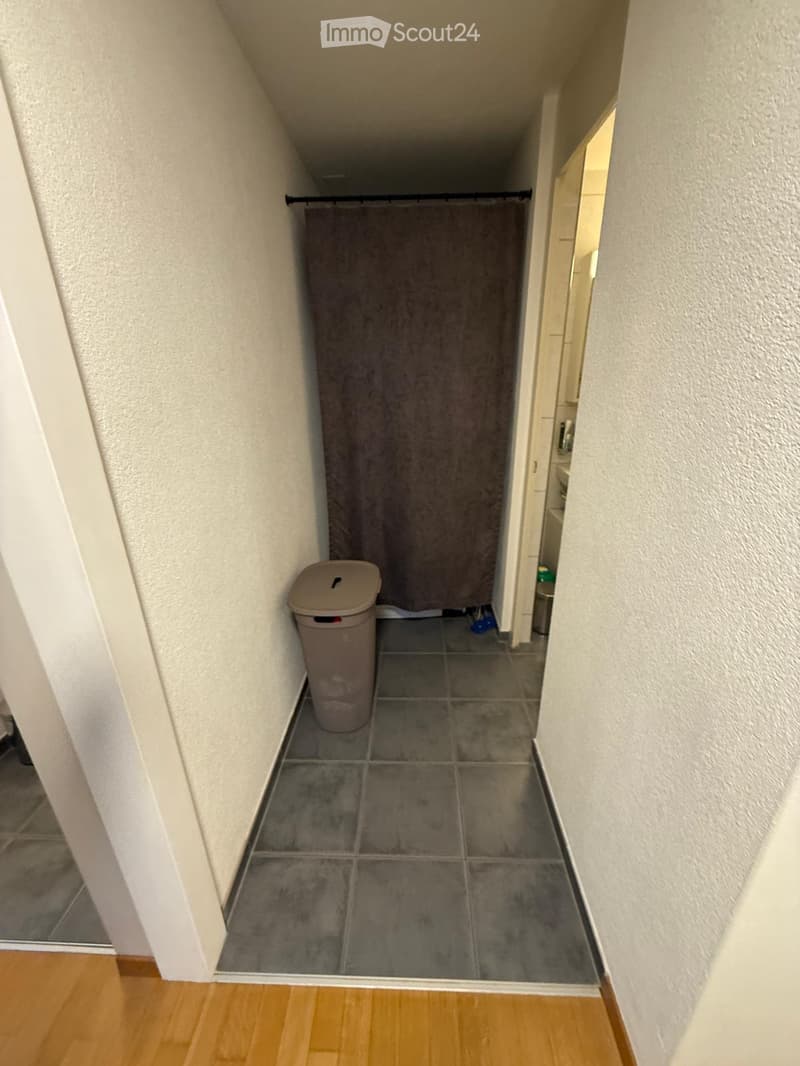 Narrow hallway with tiled floor, white walls, and a trash bin on the left side. A closet with a curtain is visible at the end of the hallway.