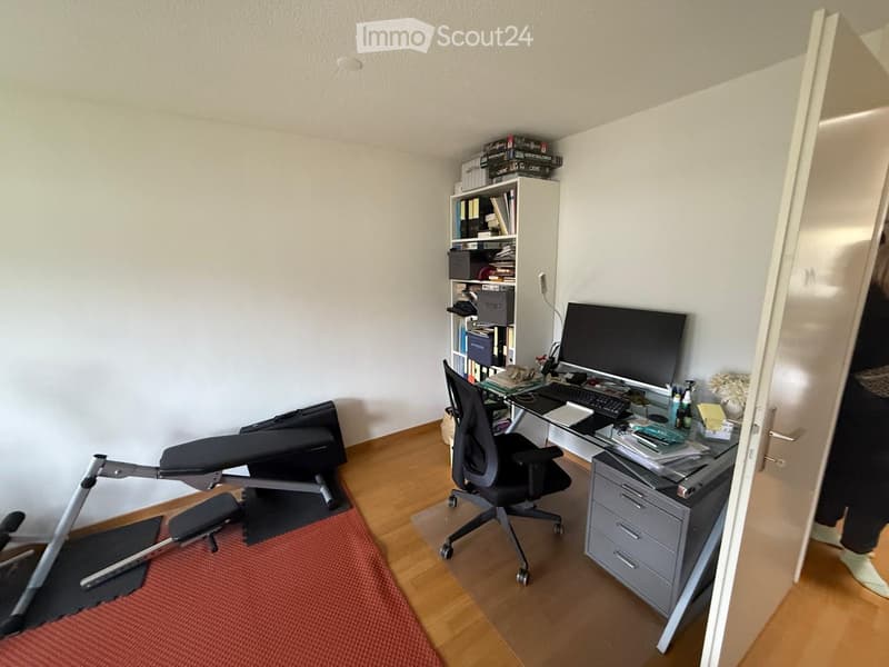 A modern, well-organized home office with a wooden floor, a large desk, a computer setup, a comfortable office chair, and a bookshelf filled with books and storage boxes.