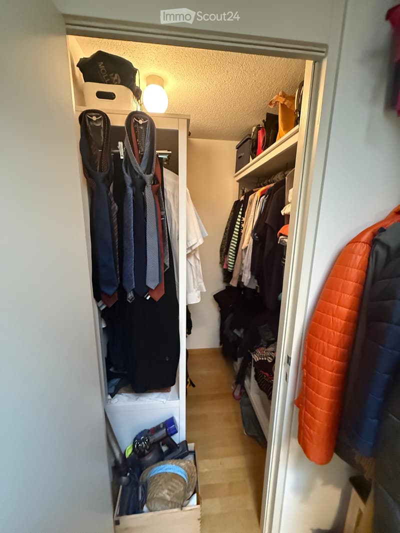 A well-organized closet with multiple sections, including hanging clothes, shelves, and storage boxes. The floor is light-colored wood, and the walls are white.