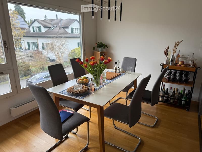 Modern dining room with a white dining table, six grey chairs, a wine rack, and a vase with tulips on the table. The room has wooden flooring and large windows with a view of the garden and neighboring house.