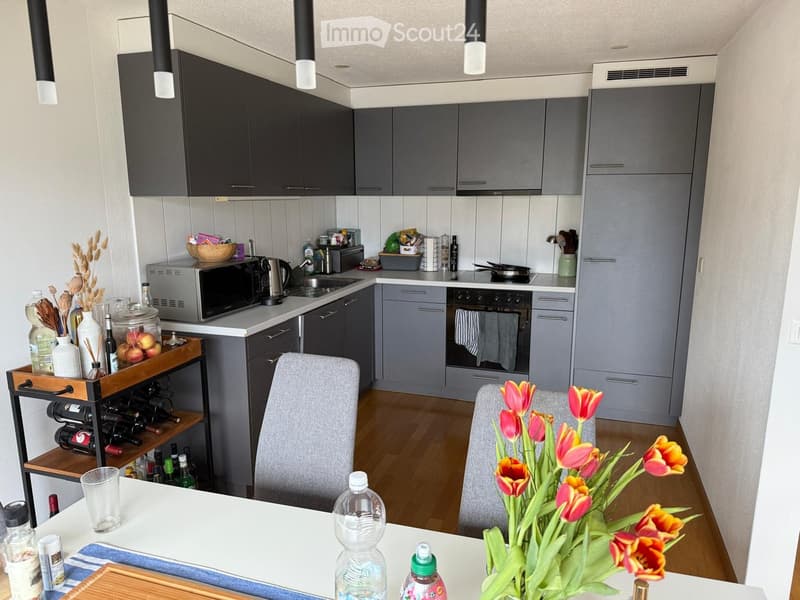 Modern kitchen with grey cabinets, stainless steel appliances, and a dining area with chairs and a table. The counter has a wine rack, a vase with tulips, and various kitchen items.