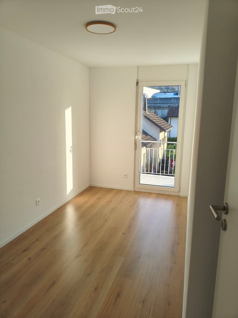 Empty hallway with light wooden flooring, white walls, and a sliding glass door leading to a balcony area.