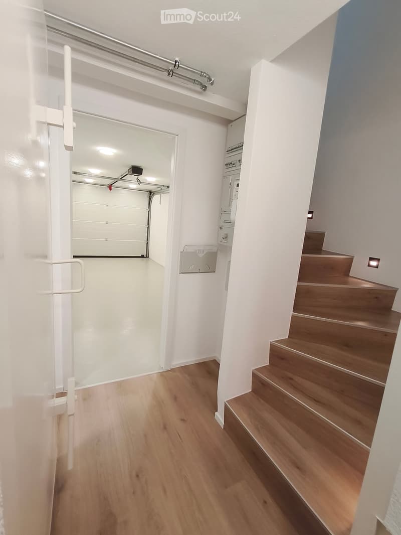 Clean, modern hallway with wooden flooring, white walls, and a staircase leading upwards. Features a white door with a handle and a small electrical panel on the wall.