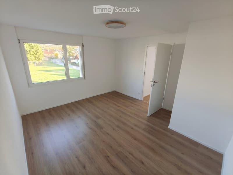 Empty room with wooden flooring, white walls, large window, and an open door leading to another room.