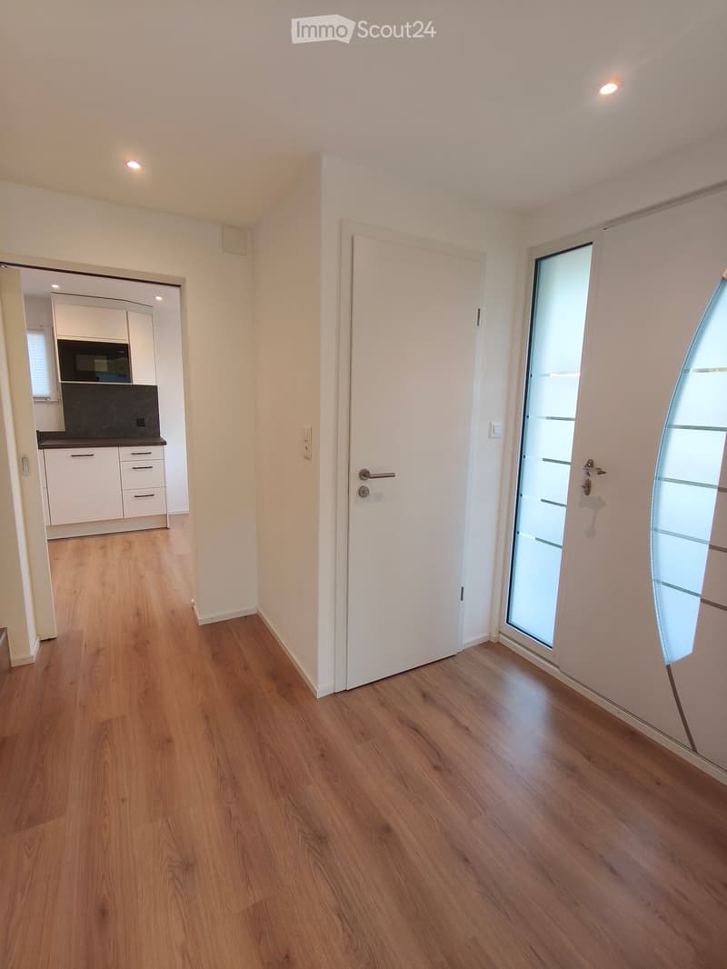 Empty hallway with wooden flooring, white walls, and recessed lighting. Features a closed white door and an open door leading to a kitchen area with white cabinets and a black countertop.