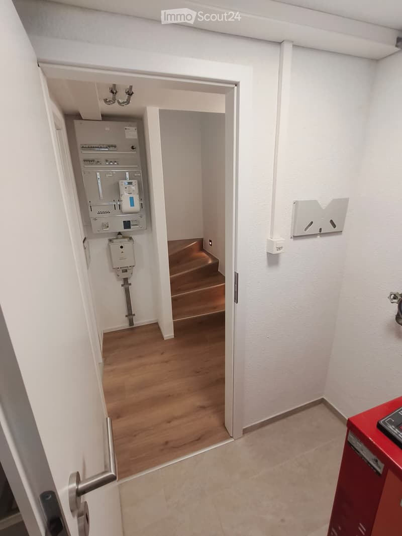 A narrow hallway with white walls and wooden flooring, featuring a door leading to another room and electrical panels visible in the background.