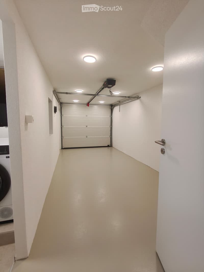 A narrow, well-lit hallway leading to a closed garage door. The walls are white, and the floor is a light beige color. There are several ceiling lights and a small camera mounted near the garage door.