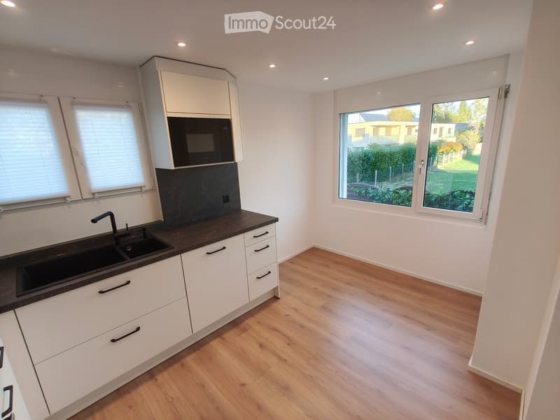 Modern kitchen with white cabinets, dark countertops, a double sink, and a microwave. The room features wooden flooring and large windows with blinds.