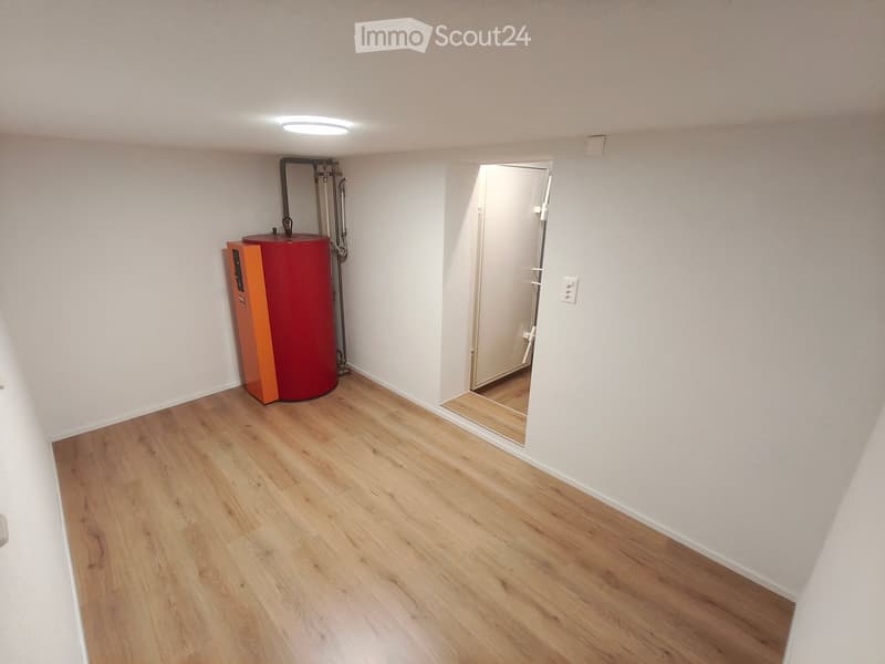 Empty room with light wooden flooring, white walls, and a red water heater in the corner. A doorway leads to another room.