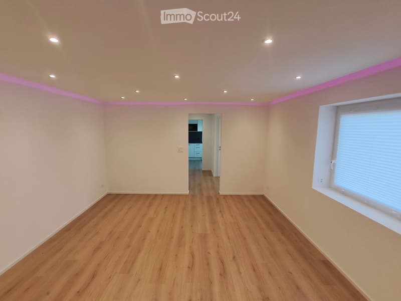 Empty room with wooden flooring, white walls, and recessed ceiling lights. A window with blinds is on the right side, and a doorway leads to another room with blue cabinets and a microwave visible in the background.