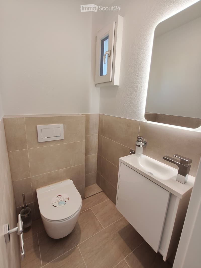 Modern bathroom with a white toilet, a white sink with a chrome faucet, a large mirror, and a small window. Beige tiles on lower walls and light-colored tiles on the floor.
