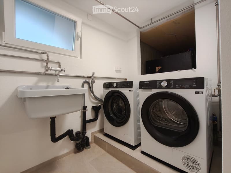 Modern bathroom with a white sink, washing machine, and dryer, featuring a window and visible plumbing.