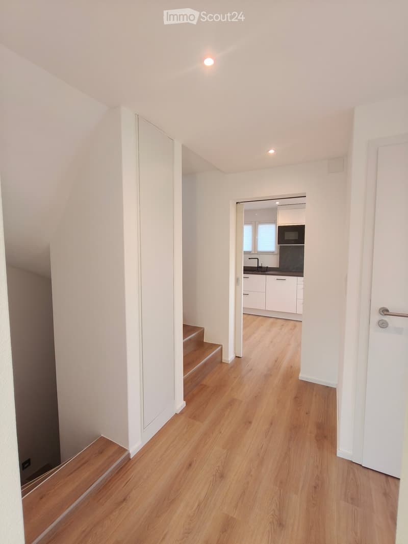 Bright hallway with wooden flooring, white walls, and recessed lighting. A staircase leads to an upper level, and an open doorway reveals a modern kitchen with white cabinets, a microwave, and a sink.