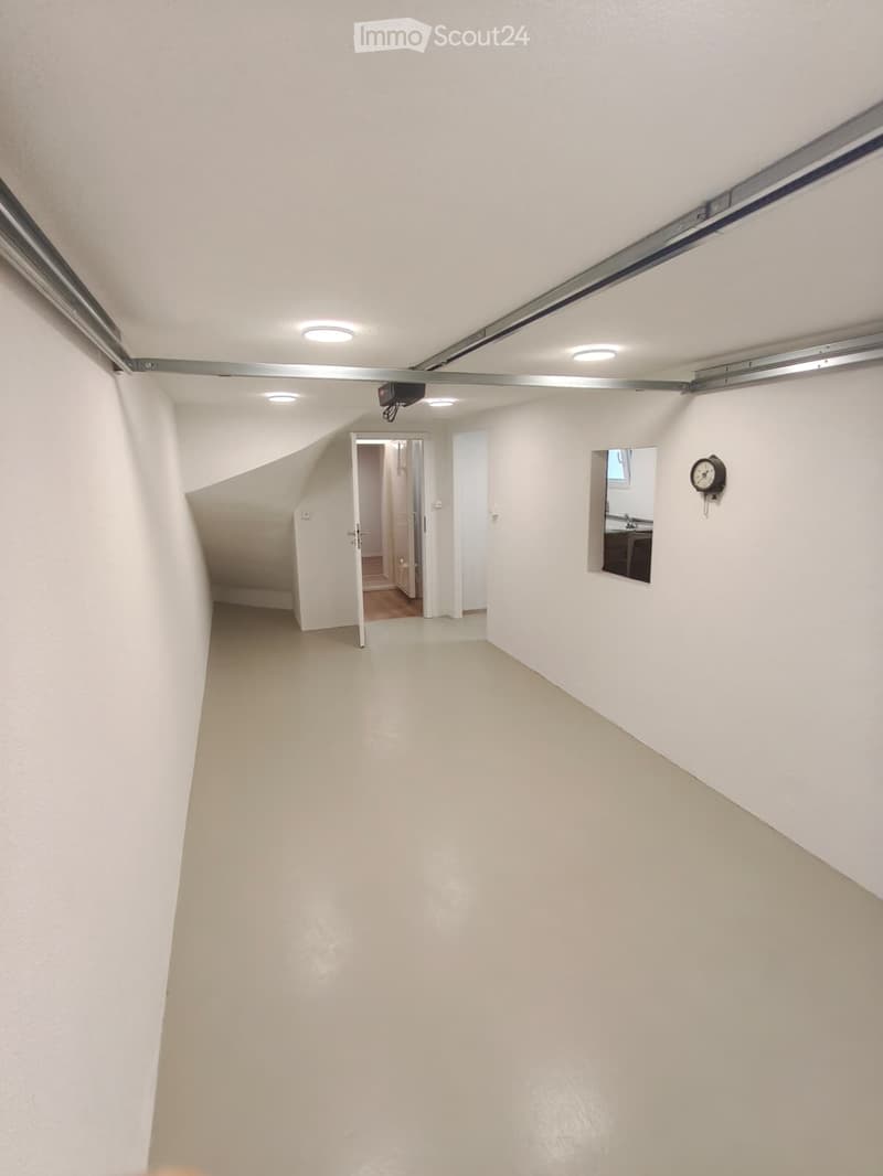 Empty hallway with white walls, a ceiling light, and an open doorway leading to another room. A small mirror and a wall-mounted clock are visible on the right side.