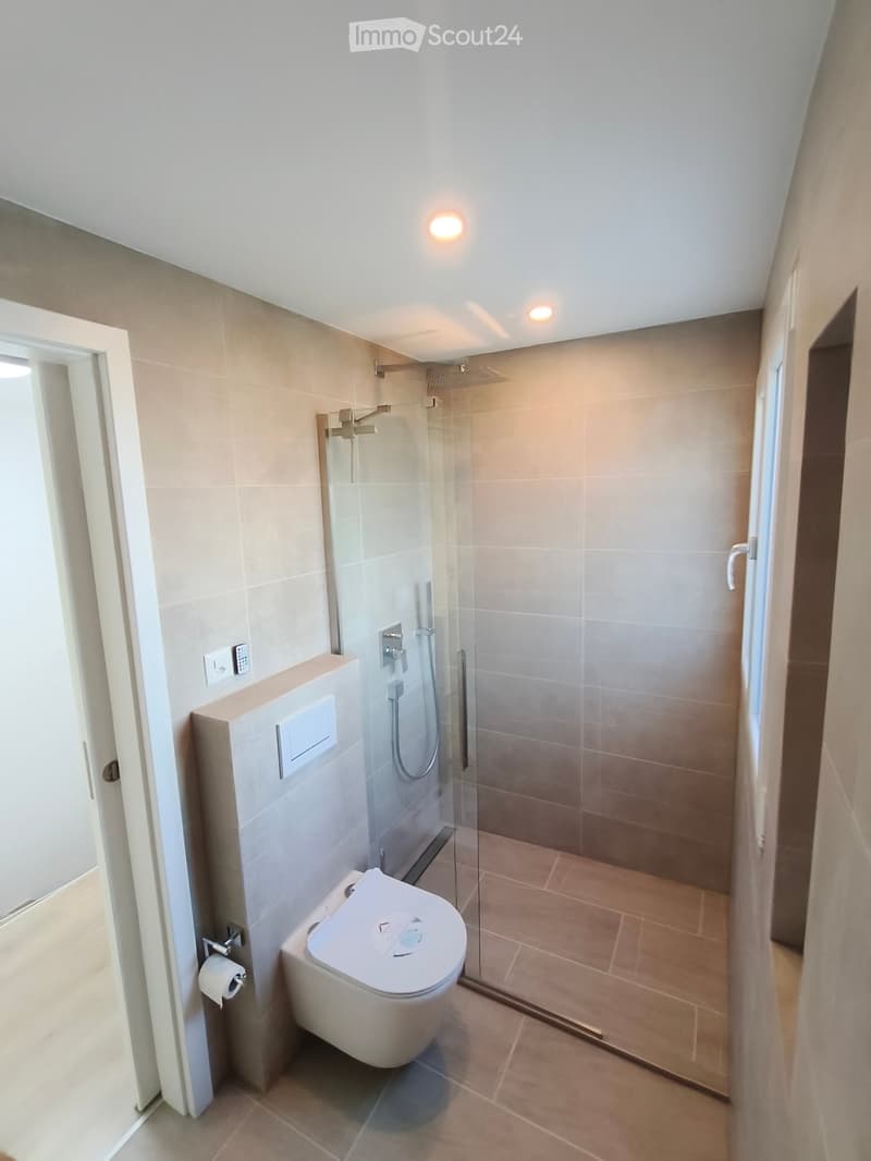 Modern bathroom with a toilet, glass-enclosed shower, and built-in toilet paper holder. Light-colored tiles and sleek fixtures.