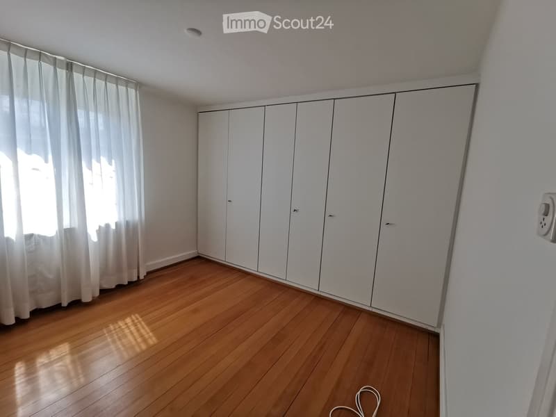 Empty room with wooden flooring, white built-in wardrobes, and sheer curtains covering a window.