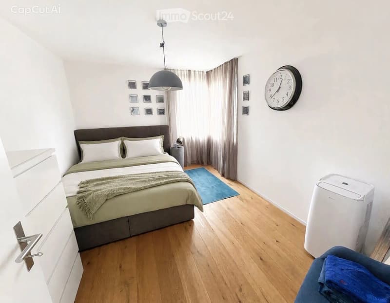 Modern bedroom with a large bed, nightstand, wall-mounted clock, air conditioner, and a small blue rug.