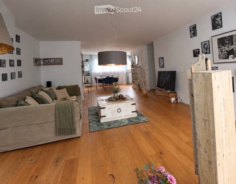 Open-plan living and dining area with wooden flooring, a beige sofa with green cushions, a coffee table, a dining table with chairs, a television, and decorative picture frames on the walls.
