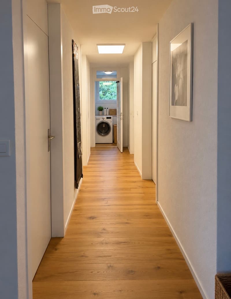 Modern hallway with light wooden flooring, white walls, and recessed ceiling lighting. A framed picture hangs on the right wall, and a small laundry area is visible at the end.