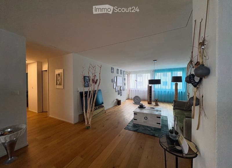 Bright living room with wooden flooring, large windows with blue curtains, a green sofa, white coffee table, floor lamp, side table, decorative branches, and picture frames on the wall.