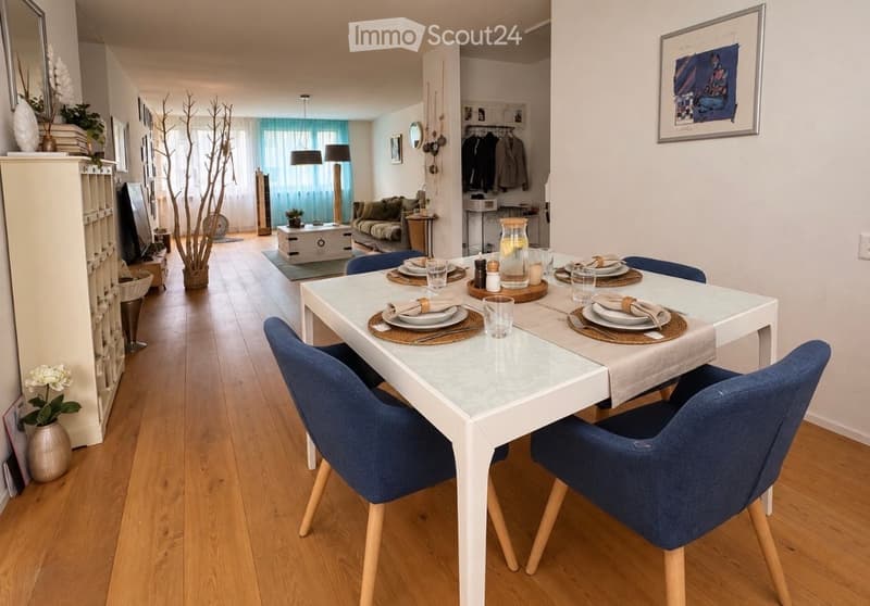 Open-plan living and dining area with wooden flooring, white dining table with blue chairs, sofa, lamps, and decorative items.