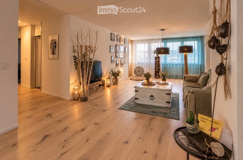 Bright living room with wooden flooring, large windows, a sofa, coffee table, television, floor lamp, and decorative items including picture frames and plants.