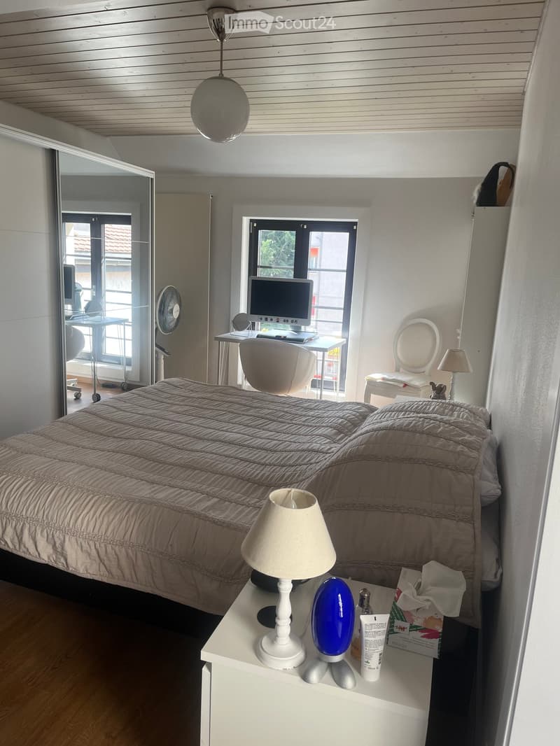 Modern bedroom with a large bed, nightstand, lamp, and a small workspace area with a computer and chair. The room features wooden flooring, white walls, and large windows with a view of the outside.