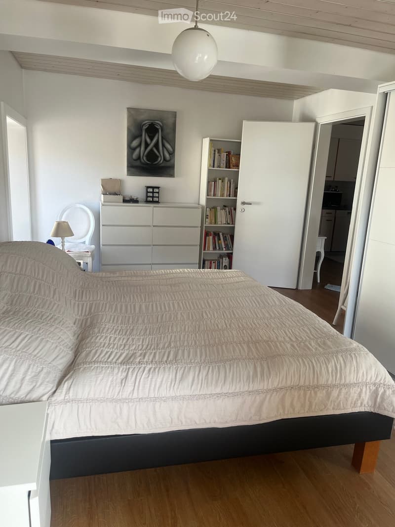 A well-lit bedroom with a large bed covered in a light-colored duvet, a nightstand with a lamp, a white chair, a cabinet, a bookshelf, and a modern artwork on the wall.