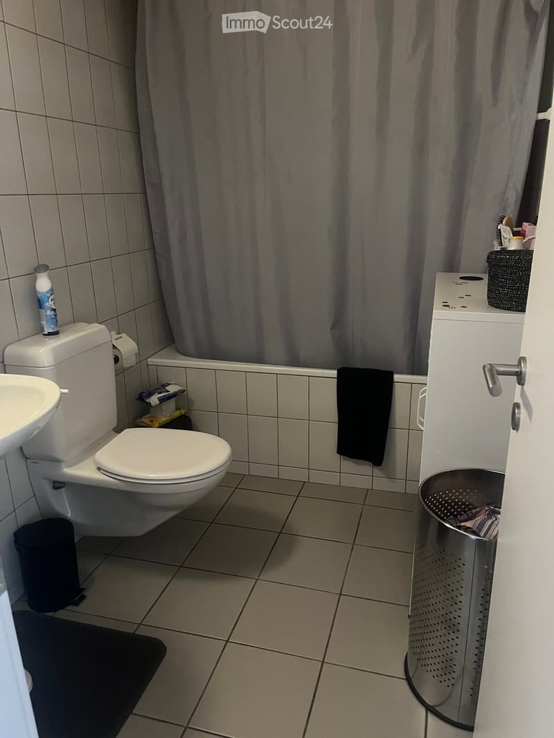 A small bathroom with a toilet, sink, shower curtain, and a towel hanging on the wall. There is a trash can and a bottle on the sink counter.