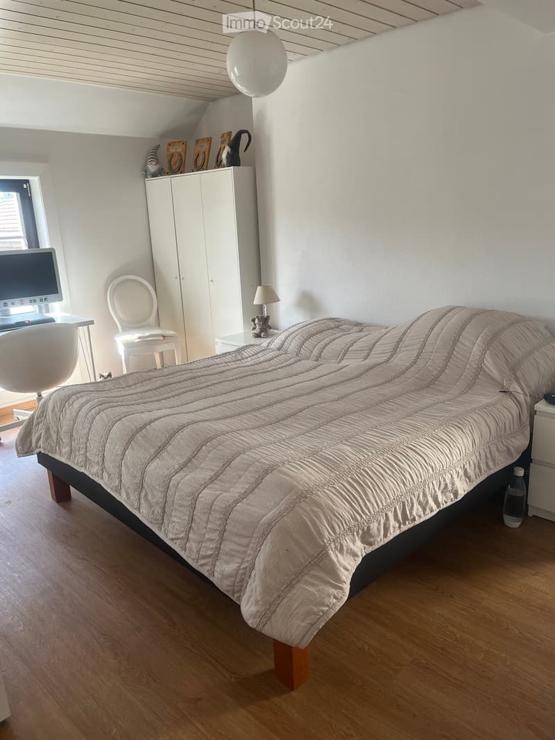 A modern bedroom with a large bed, a white chair, a small table with a lamp, a cabinet, and a computer screen on a desk.
