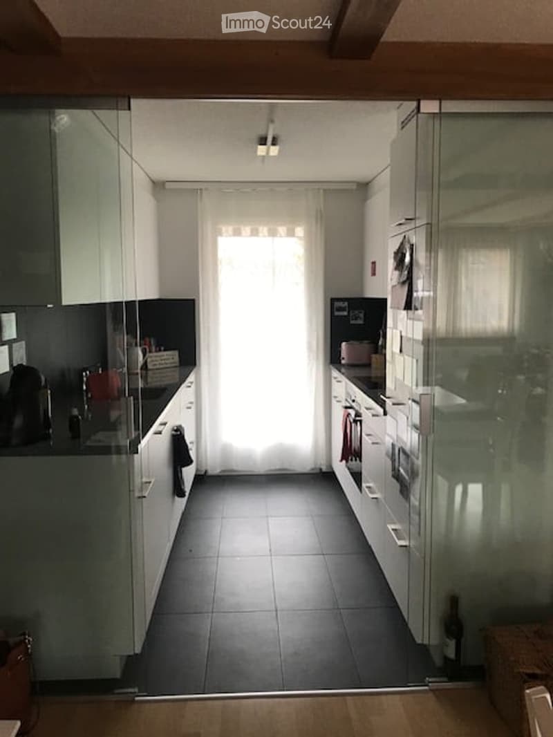 Modern kitchen with white cabinets, black tiled floor, and a large window with sheer curtains. Visible items include a countertop, sink, fridge, oven, and a wine bottle on the floor.