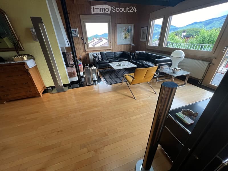 Living room with wooden flooring, large windows offering a scenic mountain view, a black leather sofa, a coffee table, a yellow chair, a side table, and a decorative mirror.
