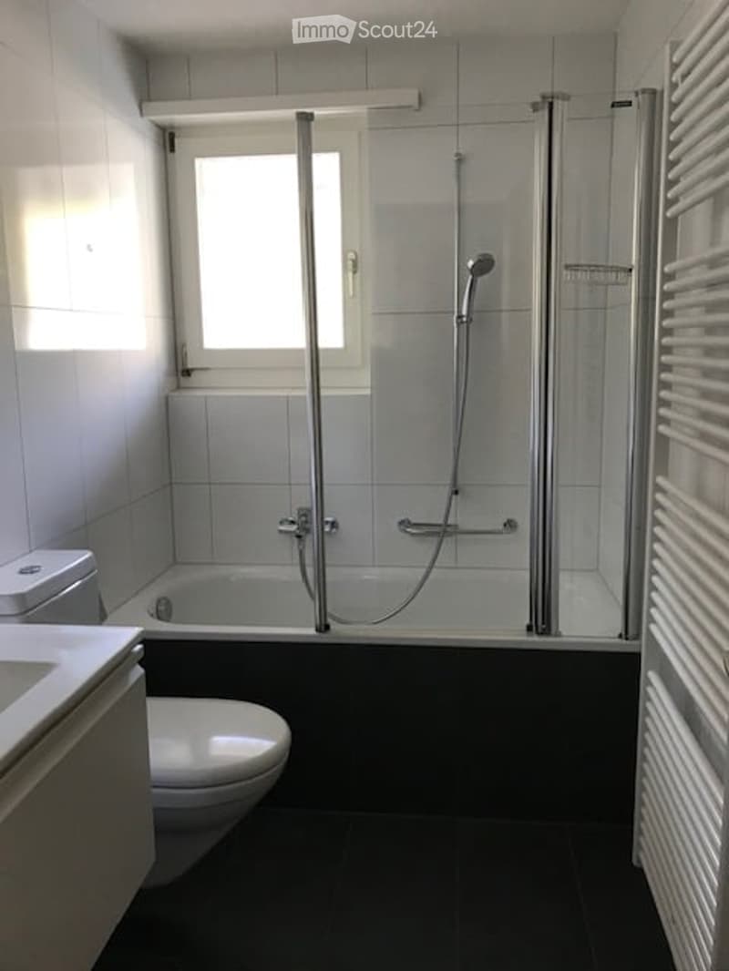 Modern bathroom with a bathtub, toilet, and a glass-enclosed shower. The space features white tiles, a window for natural light, and a white radiator on the right side.