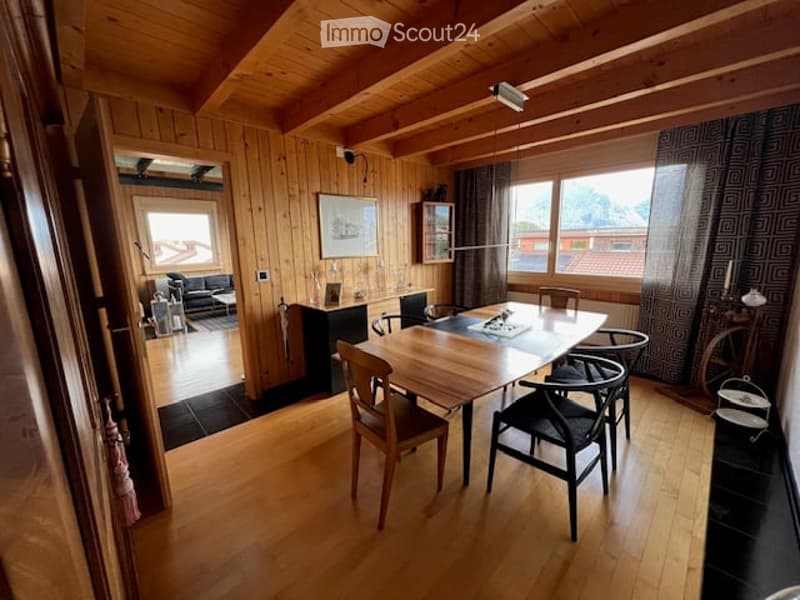 Bright dining area with wooden floors and exposed wooden beams. Features a dining table with chairs, a cabinet, and a picture frame on the wall. An adjacent living area is visible with a sofa and a table lamp.