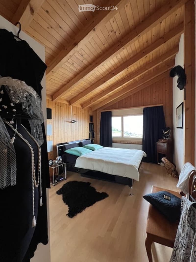 Cozy bedroom with wooden beams, a large window with dark curtains, a bed with green pillows, a nightstand, and a small dog near the window.