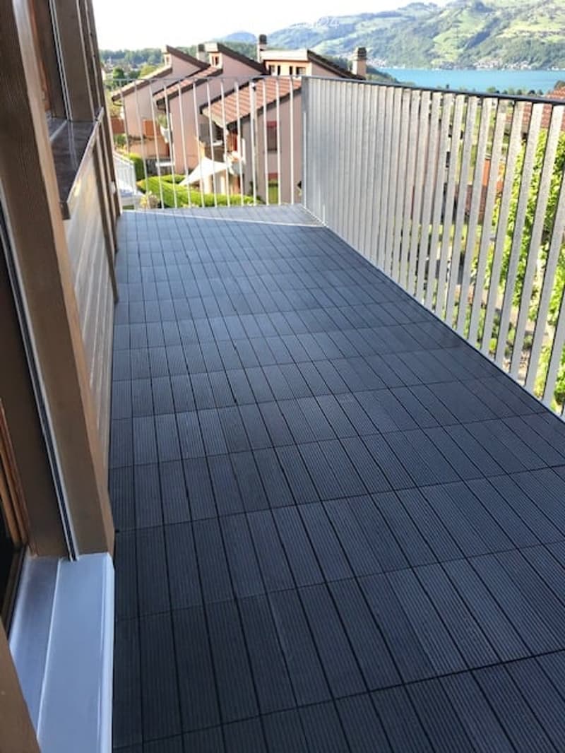 Balcony with dark grey tiled flooring and metal railing, overlooking a scenic view of houses and a lake.