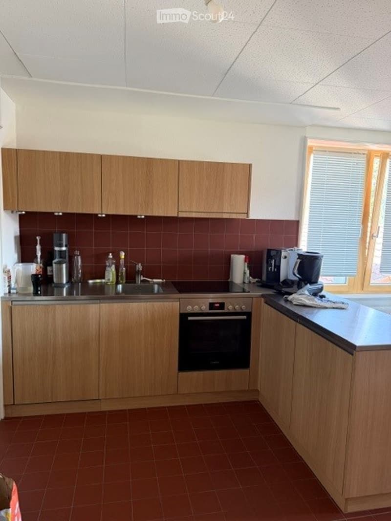 Modern kitchen with light wooden cabinets, stainless steel appliances, and a dark countertop. Features include a sink, oven, refrigerator, coffee maker, and various kitchen items on the counter.