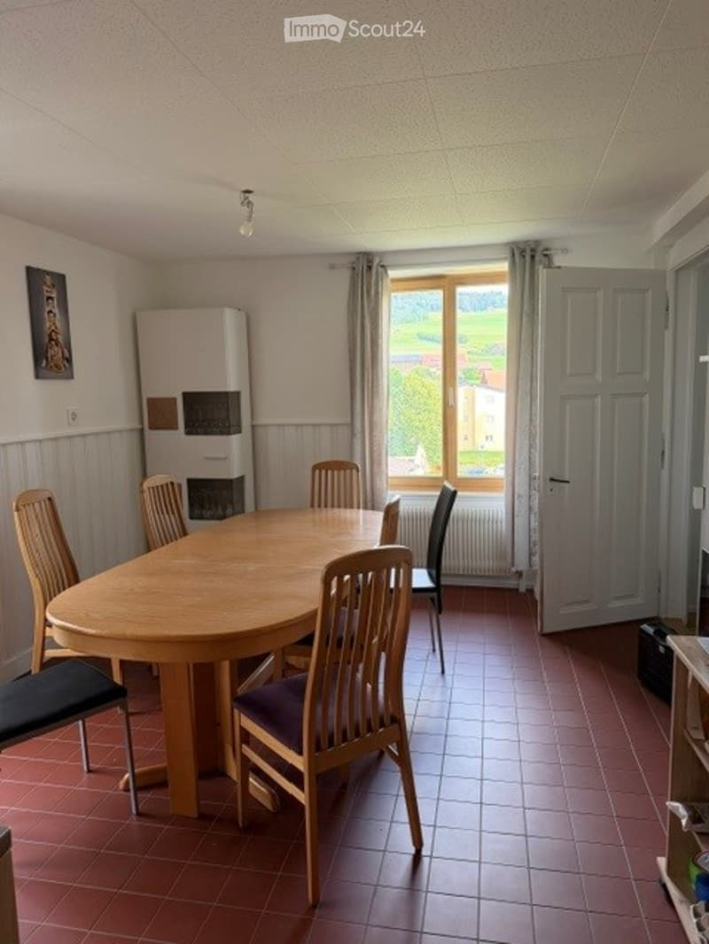 Dining room with a round wooden table surrounded by six chairs, a radiator under a large window, a white fridge, and a picture frame on the wall.