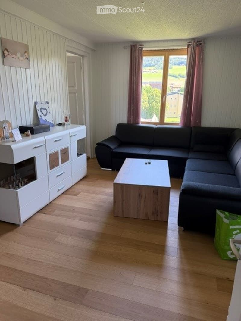 Modern living room with light wooden flooring, a black sectional sofa, a white coffee table, a white sideboard with a built-in fireplace, and a large window with pink curtains offering a scenic view.